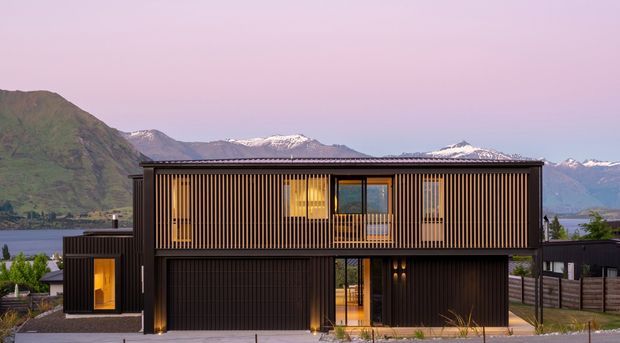 A contemporary two-storey dark-clad home viewed from the street, featuring a strong rectangular form, vertical timber screening across the upper level, large glazed openings, and warm interior lighting. A garage and entry are visible at ground level. The house sits in a mountainous lakeside setting with snow-capped peaks in the distance.