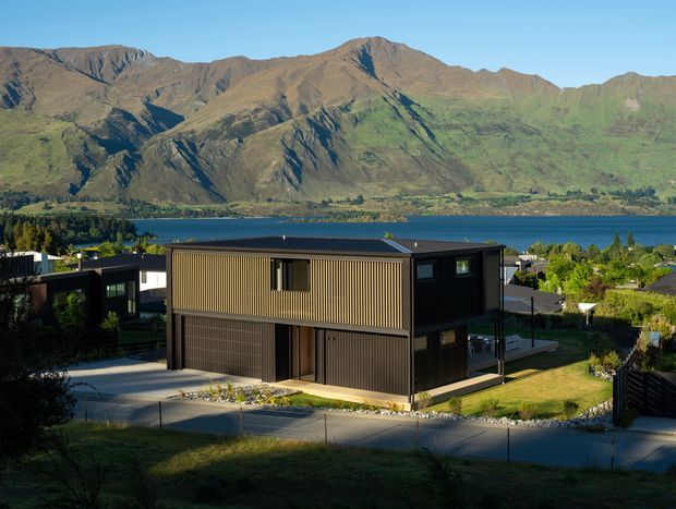 Exterior view of a contemporary two-storey dark-clad house with a boxy form, flat roof, vertical timber-look screening and large garage door. The home sits on a sloping suburban site overlooking a lake and dramatic mountainous landscape.