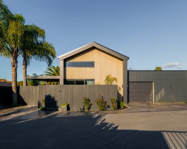 Street-facing exterior of a contemporary single-storey residence with a prominent gabled roof, warm vertical timber-clad upper facade, dark metal-framed glazing, and a large dark grey garage volume. A corrugated metal privacy fence, concrete driveway, and subtropical palms frame the house.