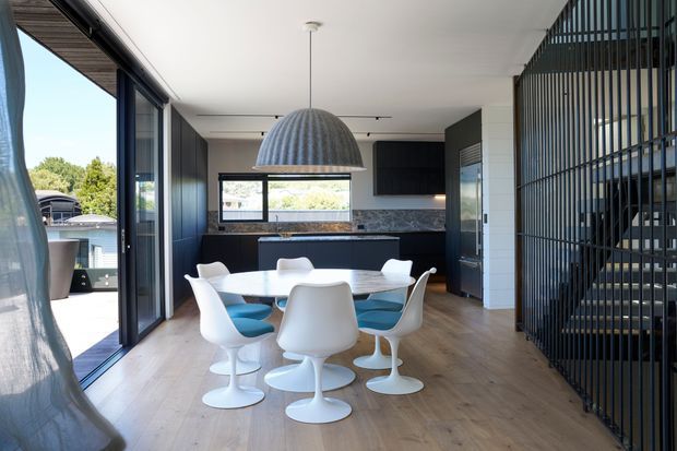 A bright contemporary open-plan dining and kitchen area with a white oval dining table and sculptural pedestal chairs, set beneath a large ribbed dome pendant light. The room has wide timber flooring, dark kitchen cabinetry, a stone benchtop and splashback, black-framed floor-to-ceiling glazing opening to the outdoors, and a black vertical-slat stair enclosure on the right.