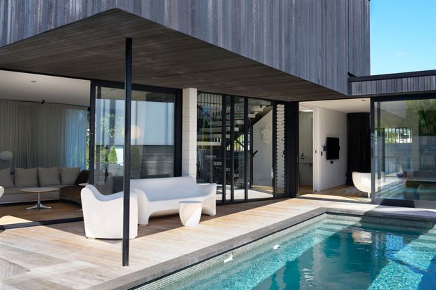 A contemporary residential courtyard viewed from the pool area, with a large swimming pool in the foreground, pale stone paving, extensive black-framed glazing, and a projecting upper volume clad in vertically textured dark timber. The open-plan living area is visible behind the glazing, along with a stairway and a covered outdoor seating zone. The scene is bright and refined, with strong indoor-outdoor connection and clean modern detailing.