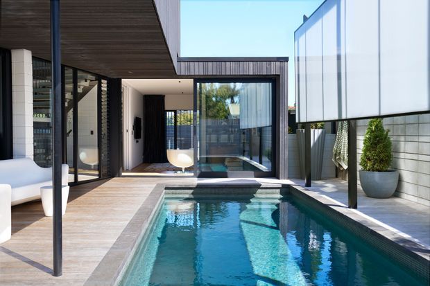 A bright contemporary courtyard viewed from beside a narrow rectangular swimming pool. The space features dark timber soffits, extensive black-framed glazing, pale timber decking, white rendered and blockwork walls, and a partially enclosed outdoor area with minimalist planting. Interior living spaces are visible through the glazing, while the pool and courtyard dominate the image.