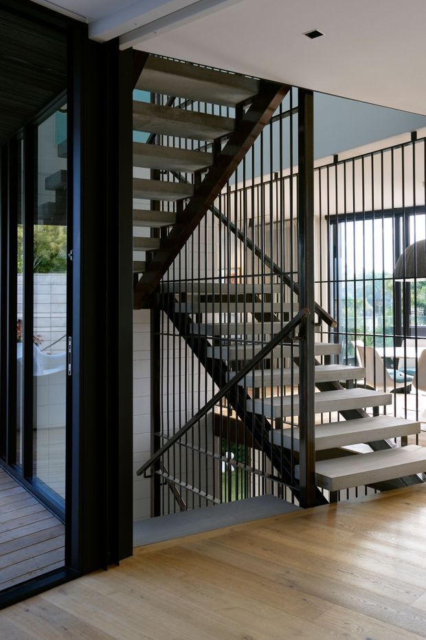 Interior view of a contemporary multi-level stairwell with open-riser timber or concrete-look steps, dark metal balustrades and structural framing. Warm timber flooring fills the foreground, while full-height glazing and an adjacent dining or living area are visible beyond the stair enclosure. The space is bright and architecturally focused, with strong vertical lines and a double-height volume.
