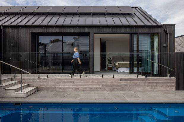 Contemporary dark-clad single-storey house facade overlooking a swimming pool. The building has a steeply pitched standing-seam metal roof, extensive black-framed glazing and sliding doors, a timber-toned terrace with glass balustrade, and a bedroom visible through the open doorway.