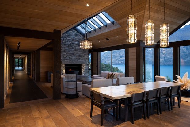 A dramatic open-plan dining and living area with warm timber lining across the ceiling and walls, dark timber-framed floor-to-ceiling glazing, and a large skylight. A substantial stone-clad fireplace forms the focal point beside a pale timber dining table with dark chairs, while the living area sits beyond. Multiple cylindrical pendant lights add a decorative glow. Through the glazing is a striking mountainous waterfront landscape.