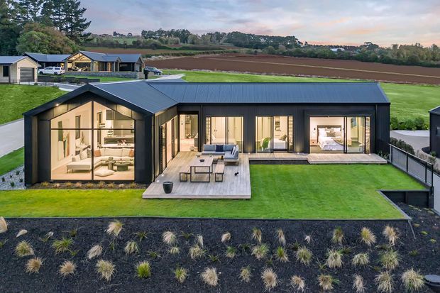 Aerial view of a contemporary single-storey home set within a landscaped rural site. The dark metal-clad house has a low pitched roof, extensive floor-to-ceiling glazing, a central timber deck with outdoor furniture, and a long rectangular swimming pool. Agricultural fields and neighboring rural buildings are visible beyond the manicured lawn.