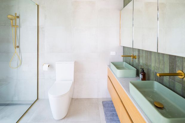 Bright contemporary bathroom with a frameless glass walk-in shower, wall-mounted gold shower fittings, a white toilet, double pale green vessel basins, and a long timber vanity with mirrored cabinetry. Large pale wall and floor tiles create a clean, light interior, while dark green vertical tiles form a feature backsplash behind the vanity.