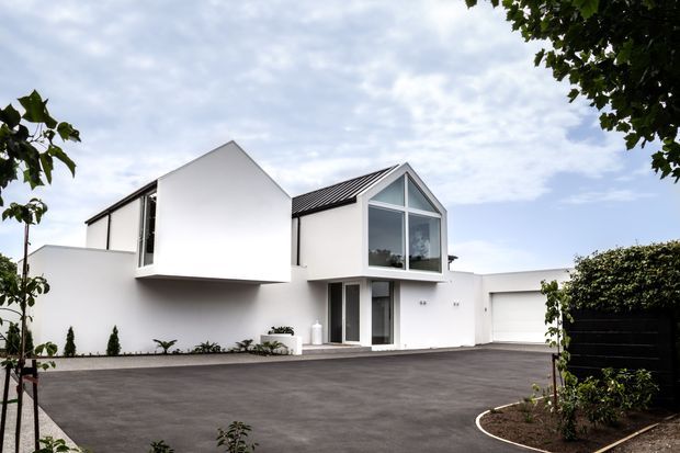 Contemporary white multi-level house with strong geometric forms, a steep gabled glazed volume, dark pitched metal roofing, and a projecting upper-floor section. The home fronts a broad dark paved driveway with minimalist planting, clipped hedges, and a partially visible garage.