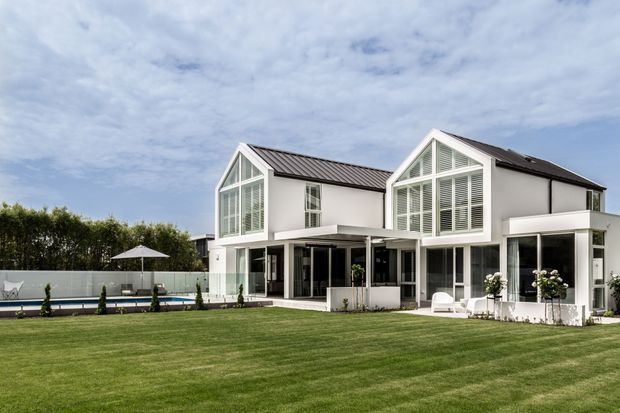Exterior view of a large contemporary white residence with two prominent pitched gable forms, dark metal roofing, extensive floor-to-ceiling glazing, and covered outdoor living areas. The home opens onto a broad manicured lawn, with a swimming pool and privacy planting visible to the side.