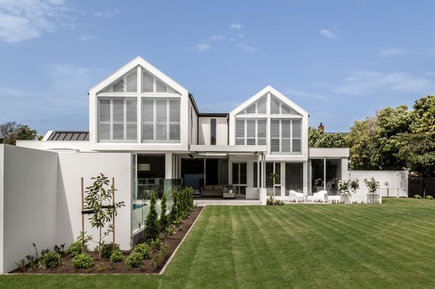 Exterior view of a refined contemporary residence with two prominent white gabled upper volumes featuring louvred glazing and decorative triangular framing. The home opens onto a broad manicured lawn and landscaped garden, with covered outdoor living areas and white outdoor furniture visible beneath the upper levels. The facade combines crisp white rendered walls, dark roof elements, extensive glazing, and strong symmetrical gable forms.