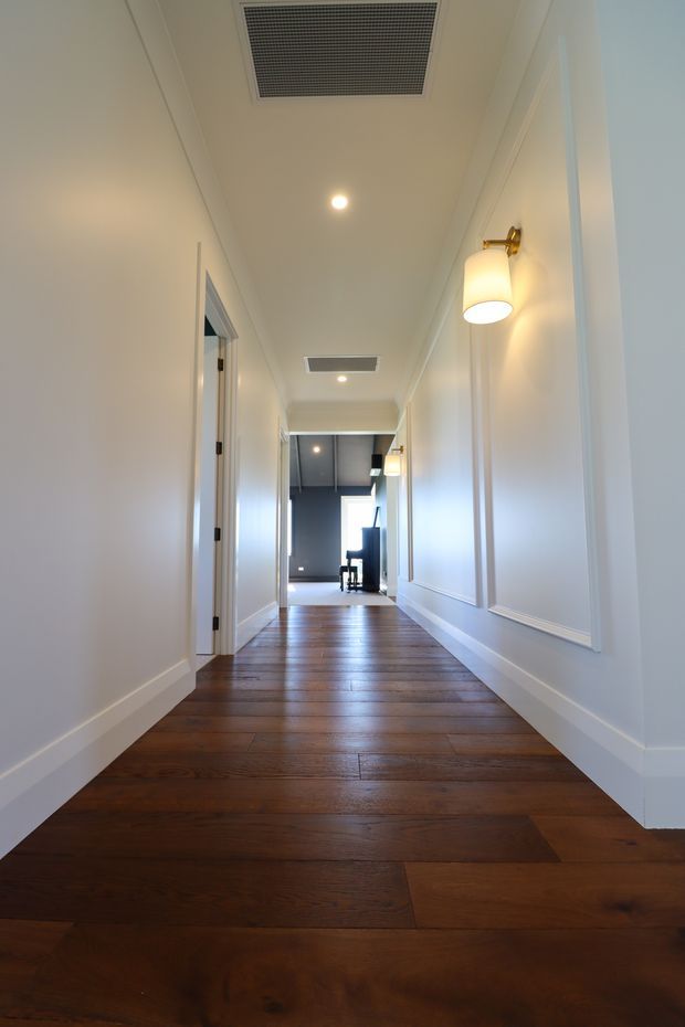 A long, narrow residential hallway with dark stained timber floorboards, white paneled walls and trim, recessed ceiling downlights, air-conditioning vents, and warm wall sconces. Multiple doorways line the corridor, which leads toward a brighter room at the far end.