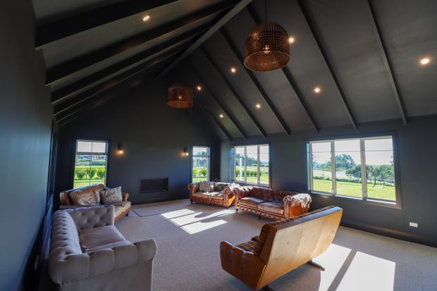 A spacious, moody living room with dark painted walls and a high pitched ceiling featuring exposed structural beams. Multiple leather sofas and armchairs are arranged around a central seating area, with large windows bringing in views of open green fields and abundant natural light. Woven pendant lights and recessed spotlights add warmth to the otherwise dark interior.