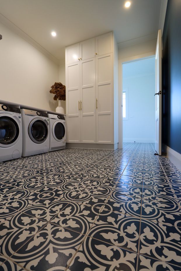 A bright, narrow laundry room with three front-loading appliances along the left wall, tall white paneled storage cabinetry at the rear, and a highly decorative patterned tile floor in dark blue and cream. The room has recessed ceiling lights, a dark blue accent wall on the right, and an open doorway leading to a pale adjoining space.