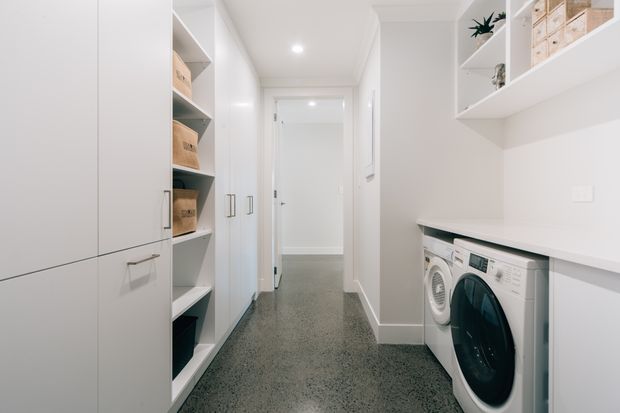 Bright, narrow contemporary laundry room with white built-in cabinetry, open shelving, storage boxes, and a concealed benchtop over front-loading washing and drying appliances. The room has a polished grey speckled floor, white walls, recessed ceiling lights, and a hallway connection at the rear.