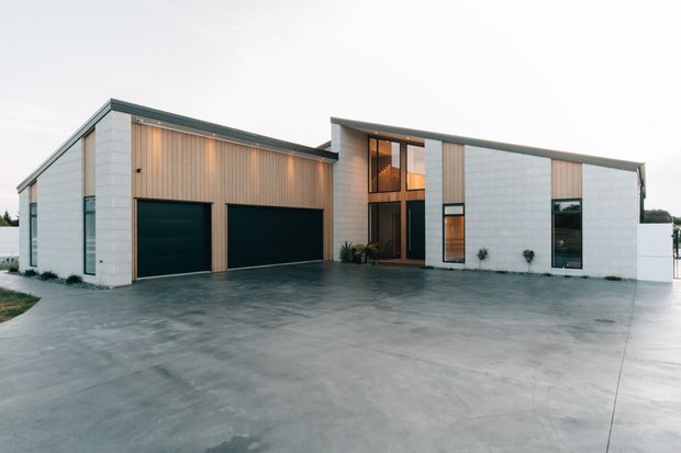 Modern single-storey residential building with a broad concrete driveway, two dark garage doors, pale white masonry or panel cladding, vertical timber-toned facade panels, and a recessed glazed entry beneath a stepped flat roofline. The clean geometric facade has large windows and minimal landscaping.