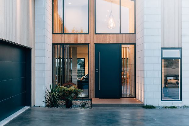 Contemporary residential facade viewed from the driveway, featuring a dark garage door, a black entry door, warm vertical timber cladding, white horizontal wall cladding, tall glazed openings, and a small planted entry bed with decorative gravel.