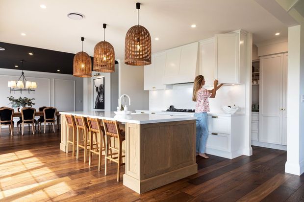 Bright open-plan kitchen and dining area with a large white island, paneled white cabinetry, dark timber flooring, and woven rattan pendant lights. A row of timber bar stools lines the island, while a formal dining table and chairs sit in the adjoining space beneath a dark ceiling feature. The room has a warm, refined residential interior with abundant natural light.