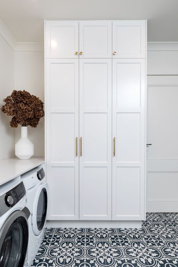 Bright, compact laundry room featuring tall white raised-panel storage cabinetry with brass knobs and pulls, white appliances along the left side, a white benchtop, and a highly decorative black-and-white patterned tile floor. The room has pale walls, simple ceiling trim, and a white ceramic vase with dried foliage.