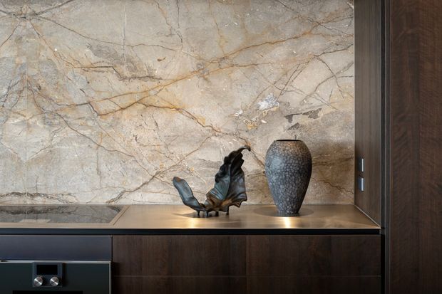 Close-up interior detail of a sophisticated built-in dark timber cabinetry unit with a warm stone or marble feature wall and matching horizontal benchtop. Sculptural metallic decorative objects sit on the bench, with integrated dark fittings visible at the lower left. The composition is refined and material-focused, with warm neutral tones and subtle lighting.