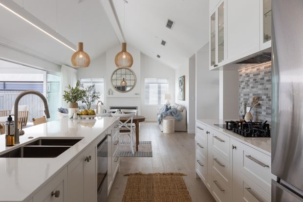 Bright open-plan kitchen with a large white island, integrated double sink, curved brushed-metal tap, white shaker-style cabinetry, and a grey mosaic tile splashback. Pendant lights hang above the island, while the space opens into a dining and living area with light timber flooring, pale walls, and abundant window light. A round mirror and soft furnishings add warmth to the otherwise clean, coastal-leaning interior.