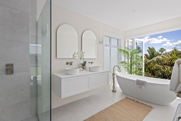 Bright, spacious contemporary bathroom with a freestanding white bathtub beside large windows, a floating double vanity with two vessel basins, arched mirrors, a glass-enclosed shower, and pale tiled surfaces. Tropical greenery and blue sky are visible outside, creating an airy resort-like atmosphere.