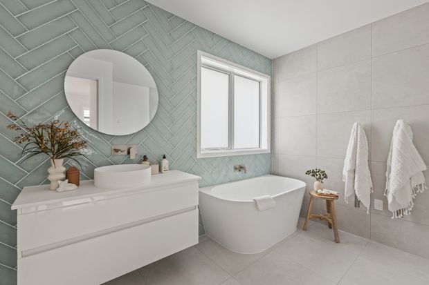 Bright contemporary bathroom featuring a floating white double-drawer vanity with a round mirror, vessel basin, freestanding white bathtub, and wall-mounted fixtures. The room combines pale grey-green herringbone tiles on the vanity and bath wall with large light-grey stone-look wall and floor tiles, white towels, and a large window.