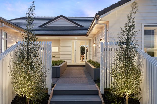 A landscaped entry courtyard leading to a light-coloured single-storey home. The approach features dark stepped decking, raised rectangular planters, illuminated olive trees, white picket-style fencing, and weatherboard-clad walls beneath dark tiled pitched roofs. A central gabled roof feature and glazed entry door are visible at the end of the walkway.