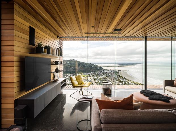 A contemporary living room with extensive floor-to-ceiling glazing overlooking a coastal bay and hillside settlement. The room features warm timber wall and ceiling lining, dark built-in media cabinetry, a large sectional sofa, sculptural coffee tables, and a mustard lounge chair. The space has a refined, warm modern character with a strong indoor-outdoor connection and expansive coastal outlook.