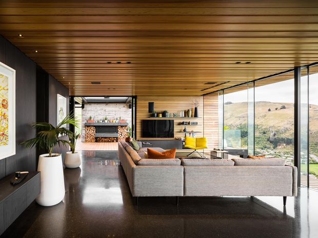 A contemporary open-plan living room with a large grey sectional sofa facing a wall-mounted television and built-in shelving. The room features a warm timber slatted ceiling, dark charcoal wall panelling and cabinetry, polished dark flooring, expansive floor-to-ceiling glazing, and a bright yellow occasional chair. A fireplace with stacked firewood is visible at the rear, while the glazing overlooks a dry rolling landscape.