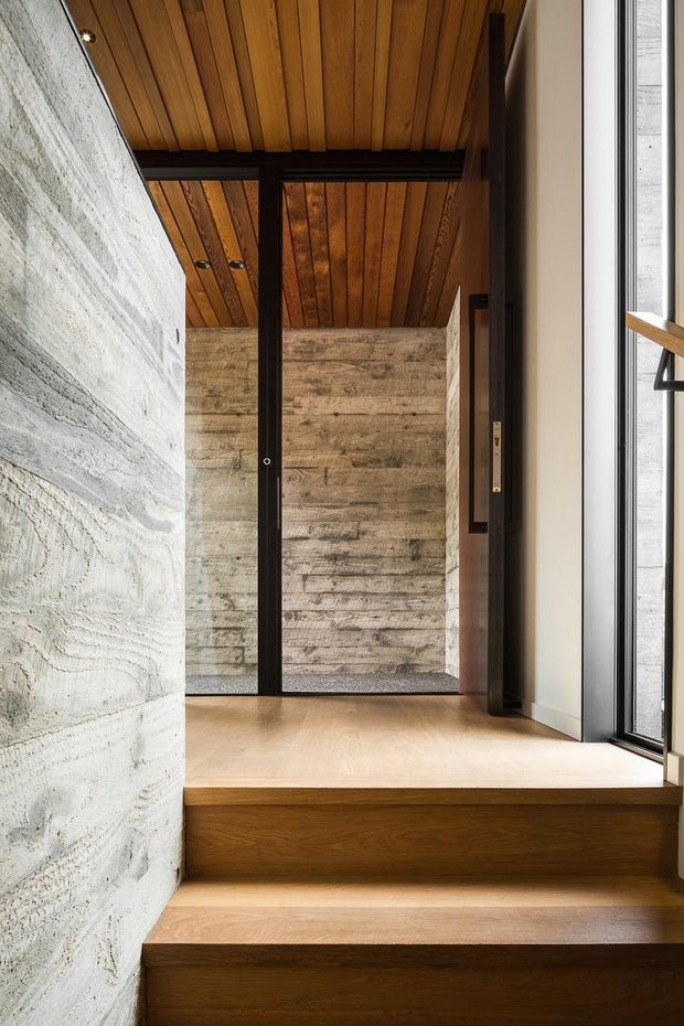 A carefully detailed interior entry or stair hall with broad solid timber steps and floor, a warm timber-planked ceiling, dark metal-framed glazing and doors, and textured pale masonry walls. The view rises toward a small landing and an open doorway, creating a layered, tactile contemporary architectural composition.