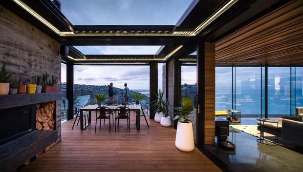 A sophisticated open-plan indoor-outdoor living and dining area with extensive floor-to-ceiling glazing overlooking a coastal urban landscape and water. The space features warm timber decking and ceiling battens, a dark framed steel or aluminium structure, a stone fireplace wall with integrated firewood storage, a dining table, potted plants, and a lounge area beyond. Large retractable or open glazed sections create a strong connection to the view, while linear integrated lighting adds a refined contemporary character.