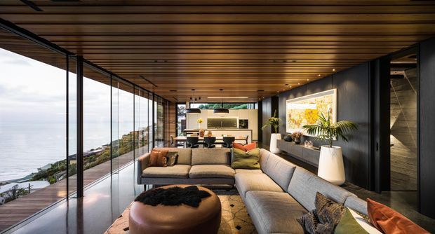 A refined contemporary living room with a large sectional sofa and round ottoman, opening through floor-to-ceiling frameless glazing to a coastal landscape. Warm timber ceiling battens, dark feature walls, integrated shelving, indoor planting and artwork create a sophisticated indoor-outdoor setting.