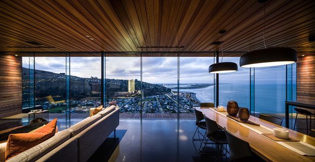 A dramatic contemporary open-plan living and dining space with full-height glazed walls overlooking a coastal bay and dense hillside settlement. The room features a warm timber-lined ceiling, dark polished flooring, a large neutral sectional sofa, and a long timber dining or bar counter with pendant lighting. The expansive glazing and panoramic elevated outlook dominate the composition.