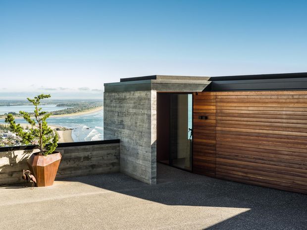 A modern rooftop terrace or deck overlooking a coastal bay and beach. The scene features a low, flat-roofed structure with a raw concrete wall and entry volume, horizontal timber cladding, dark metal roof edging, and a glazed door. A concrete parapet, potted tree, and broad paved terrace occupy the foreground, with expansive water views beyond.