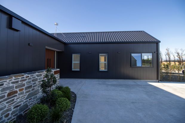 A contemporary single-storey dark-clad building exterior with a low-pitched corrugated metal roof, simple rectangular windows, and a sheltered timber-toned entry. A large smooth concrete driveway or courtyard occupies the foreground, bordered by a low natural-stone retaining wall and clipped landscaping.