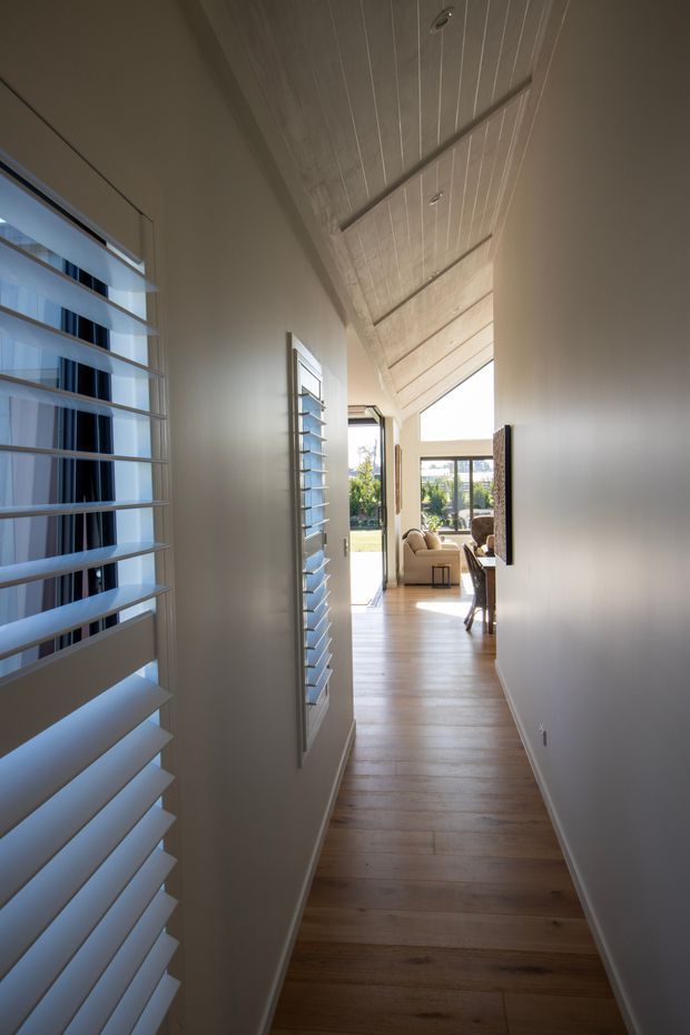 A narrow, bright residential hallway with warm timber flooring, smooth pale walls, and a vaulted white timber-panelled ceiling with recessed downlights. Full-height louvred windows and doors line the left side, bringing in daylight and providing views toward a living area and garden beyond. A neutral-toned sofa and dining furnishings are partially visible at the end of the hall.