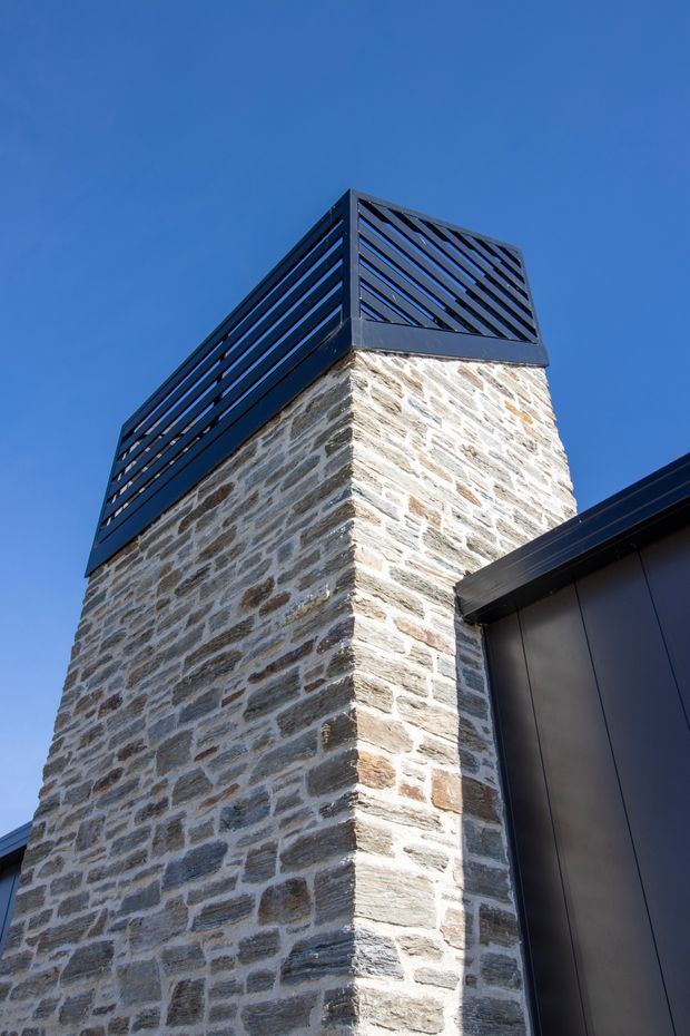 Close-up upward view of a contemporary building exterior featuring a tall chimney or tower clad in irregular light and mid-brown natural stone masonry. The upper section has a dark metal louvred cap or screen, while adjacent roof and wall surfaces use dark modern metal cladding. The image is set against a clear blue sky.