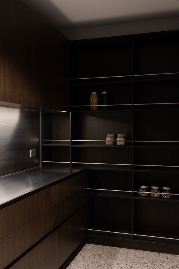 A dark, sophisticated built-in pantry or kitchen storage area with extensive charcoal-black cabinetry and open shelving. The cabinetry has a deep dark timber finish, slim horizontal metal shelf edges, and integrated drawers. A stainless-steel benchtop and matching splashback occupy the left side, while jars and pantry items sit on the shelves. The space is softly lit and grounded by a speckled terrazzo-like floor.