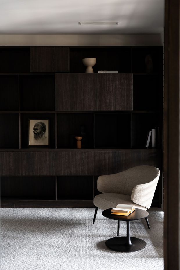A moody, refined reading nook featuring a dark built-in timber storage wall with open shelving, drawers and cabinetry, styled with minimal ceramics, books and artwork. A sculptural upholstered lounge chair and small black side table sit on a pale textured carpet, with strong sunlight creating contrast across the room.