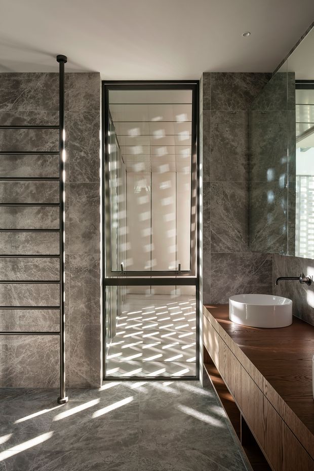 Contemporary bathroom with large grey marble-look wall and floor tiles, a dark timber floating vanity with a white vessel basin, black wall-mounted tapware, and a frameless mirror. A narrow full-height glazed opening brings in strong patterned sunlight, while a slim black heated towel rail adds a functional vertical element. The space has a refined, warm-modern character with high contrast between dark stone surfaces, timber joinery, and bright natural light.