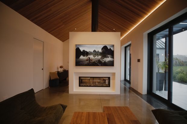 Warm contemporary living room with a dramatic pitched timber-lined ceiling, exposed dark central beam, polished concrete floor, and a minimalist white fireplace wall with a built-in firebox and landscape artwork. Large black-framed glazed doors open toward a bright outdoor view, while dark low-profile seating and timber coffee tables occupy the foreground.