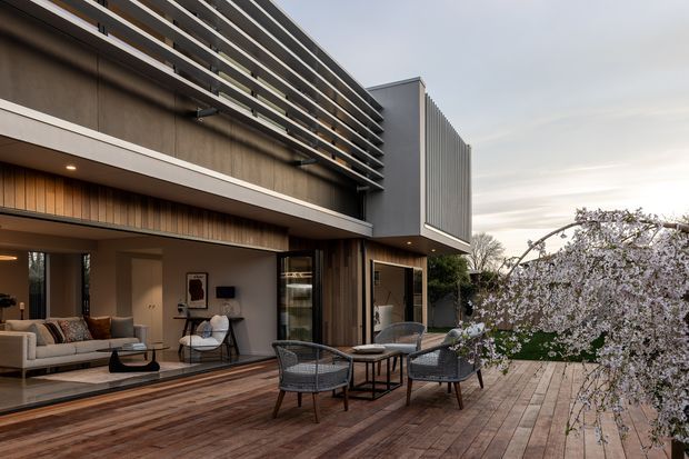 Contemporary multi-level residential facade opening onto a broad timber deck and outdoor seating area. The building combines dark vertical timber cladding, smooth grey wall surfaces, extensive glazing, and prominent horizontal privacy or sun-shading slats. A living room is visible through the open glazing, while the main focus is the exterior terrace and architectural envelope. The scene has a refined, warm contemporary character with landscaped planting at the deck edge.
