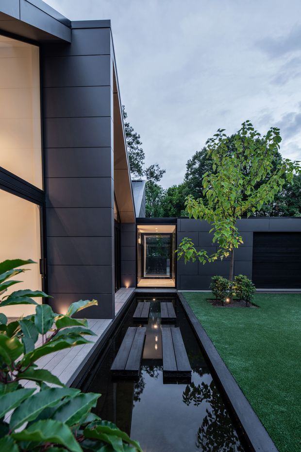 A contemporary dark-clad residential courtyard viewed from within the landscaped outdoor space. The narrow courtyard features a long reflecting pool with stepping platforms, manicured lawn, small trees and shrubs, dark linear cladding, large glazed openings, and warm architectural lighting. The composition emphasizes a calm, private modern garden and water feature.