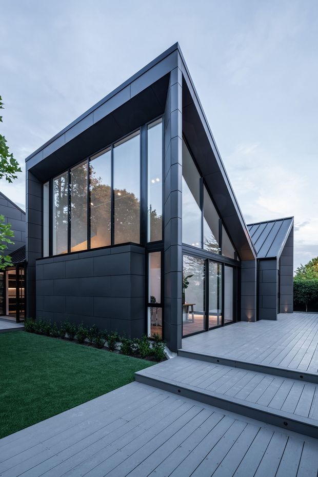 Exterior view of a contemporary, angular dark-clad house with a steeply pitched roof, extensive floor-to-ceiling glazing, and a broad multi-level timber-look deck with steps. The scene shows a landscaped lawn and hedging around the building, with a cool dusk atmosphere and strong geometric roof and facade lines.