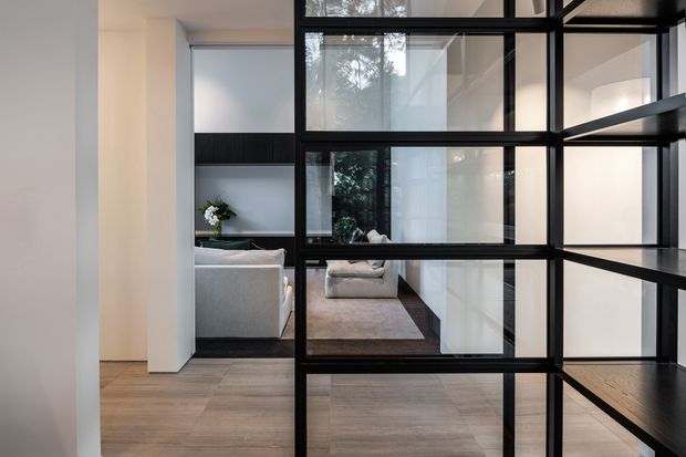 Interior view through a black metal-framed glazed partition into a contemporary living room. The space features pale timber flooring, white walls, a grey sofa, a dark built-in wall unit, and a large neutral rug. Reflections and views through the glass add depth, while the strong black grid framing creates a graphic architectural feature.