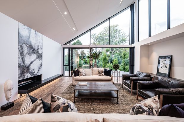 A spacious contemporary living room with a dramatic double-height glazed wall overlooking greenery. The room features a pitched ceiling with recessed linear lighting, dark timber flooring, a large textured area rug, a marble-look fireplace wall, a neutral sectional sofa, black leather lounge chairs, and a substantial dark timber coffee table. Minimal artwork and sculptural decor complete the refined interior.