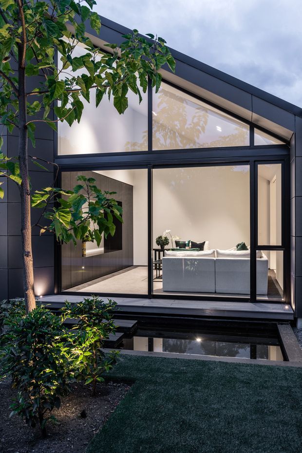 Exterior view of a sleek contemporary residence framed by dark cladding and extensive floor-to-ceiling glazing. The illuminated living room and white sofa are visible through the windows, while a landscaped garden with a reflecting pool sits in the foreground. The composition highlights the strong indoor-outdoor connection, clean geometric lines, and dark monochromatic facade.