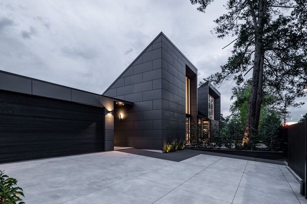 A contemporary residential exterior photographed at dusk, featuring angular gabled volumes clad in dark rectangular panels, a broad integrated garage, tall warm-lit vertical glazing, and a large pale paved driveway. Mature trees and dark boundary fencing frame the home.