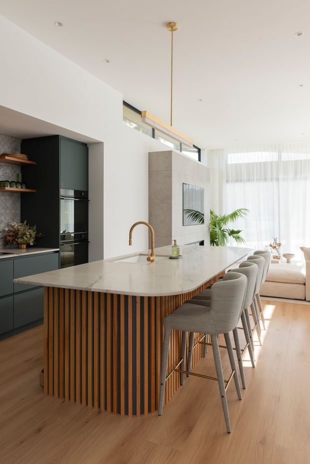 Bright open-plan kitchen and living area centred on a large island with a white stone benchtop, integrated sink and curved brass tap. The island features a striking vertical timber slat base with dark recessed accents and seating for four upholstered bar stools. Matte dark cabinetry, integrated appliances and open shelving sit against the left wall, while a tiled fireplace or feature partition, sheer curtains, indoor plants and a pale timber floor extend into the living area. A linear brass pendant hangs above the island.
