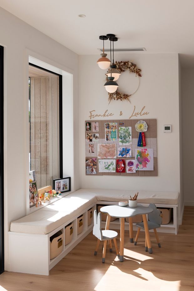 A bright, warm children's activity and dining nook with an L-shaped built-in bench and open cubby storage beneath upholstered seats. A small round children's table with matching chairs sits in front of a wall-mounted art display board labelled “Frankie & Lulu,” while three pendant lights hang above. The room has pale walls, warm timber flooring, a large window, and playful artwork and craft displays.